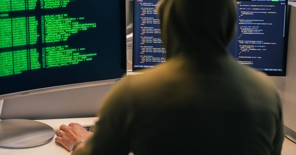 A person in a hoodie coding on dual monitors, depicting cybersecurity and hacking themes.