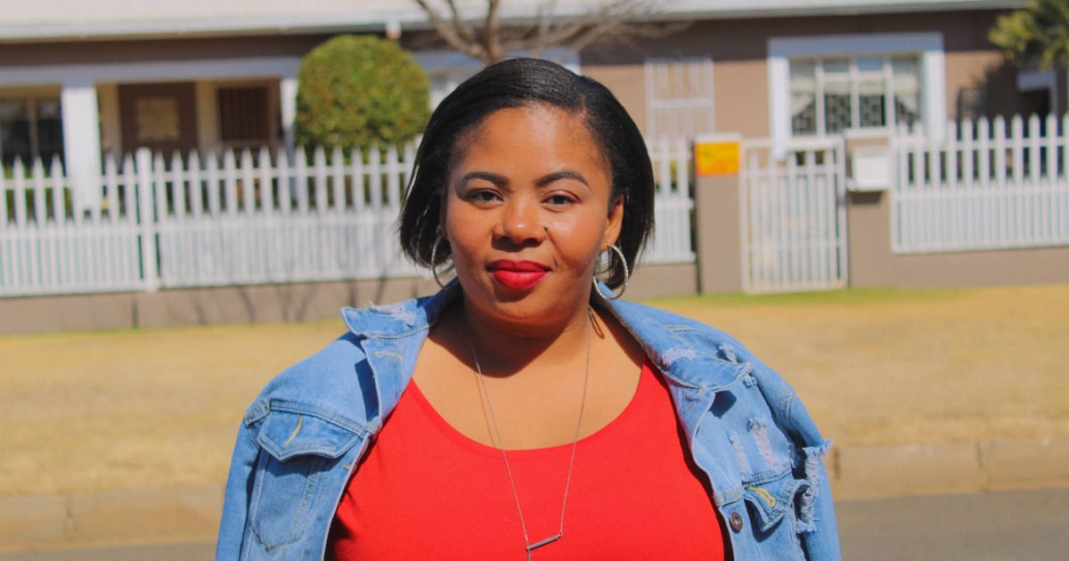 African woman in a red dress and denim jacket confidently holding her smartphone outdoors.