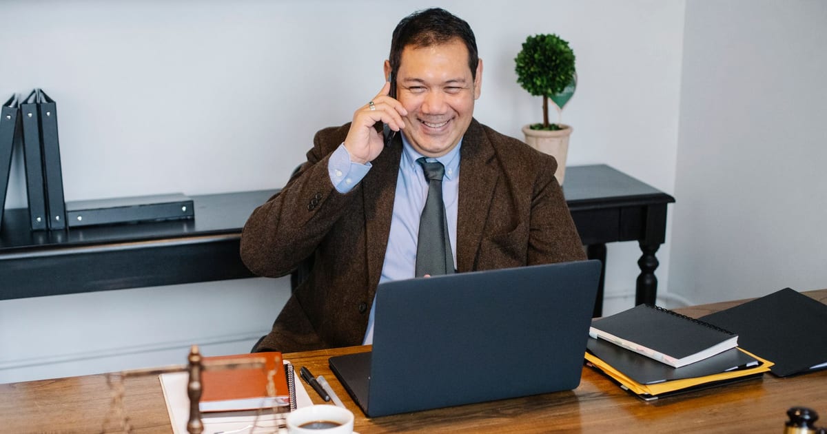 Joyful Asian male lawyer wearing formal suit having conversation on mobile phone and using laptop while working in modern law firm