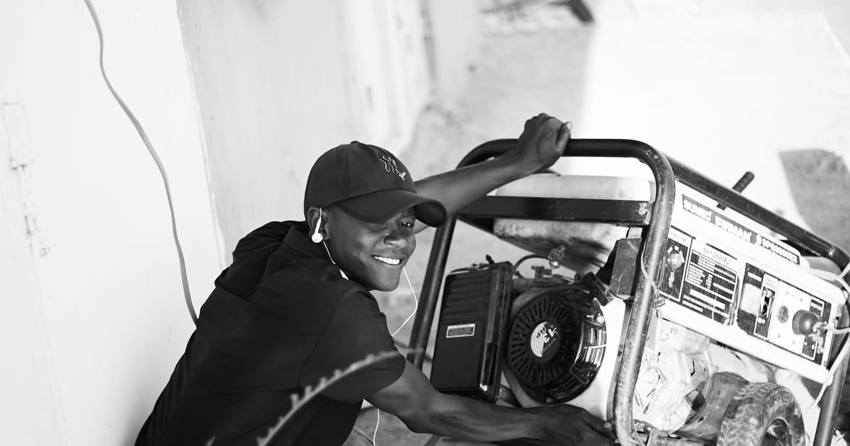 A cheerful young man wearing a cap and earphones operating a portable generator outside.