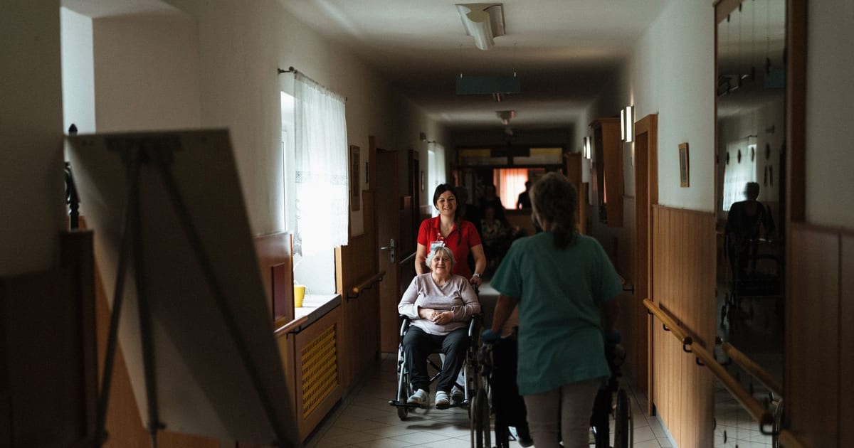 Caregiver assisting elderly woman in a wheelchair through a retirement home corridor.