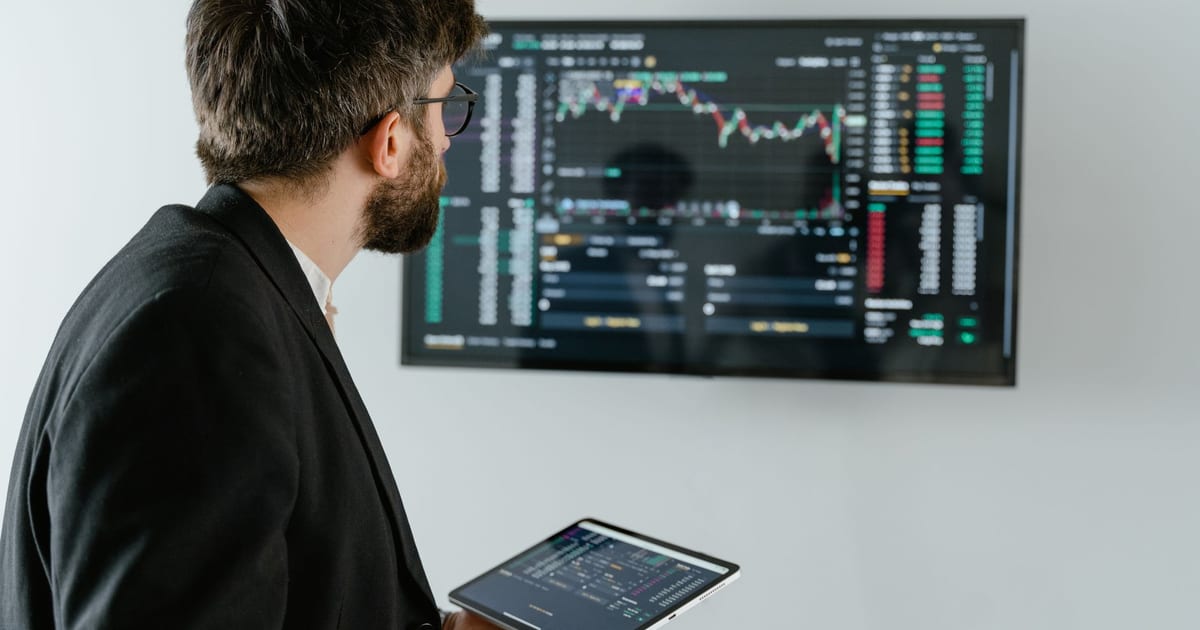 A businessman examines stock market data displayed on a monitor, holding a tablet.