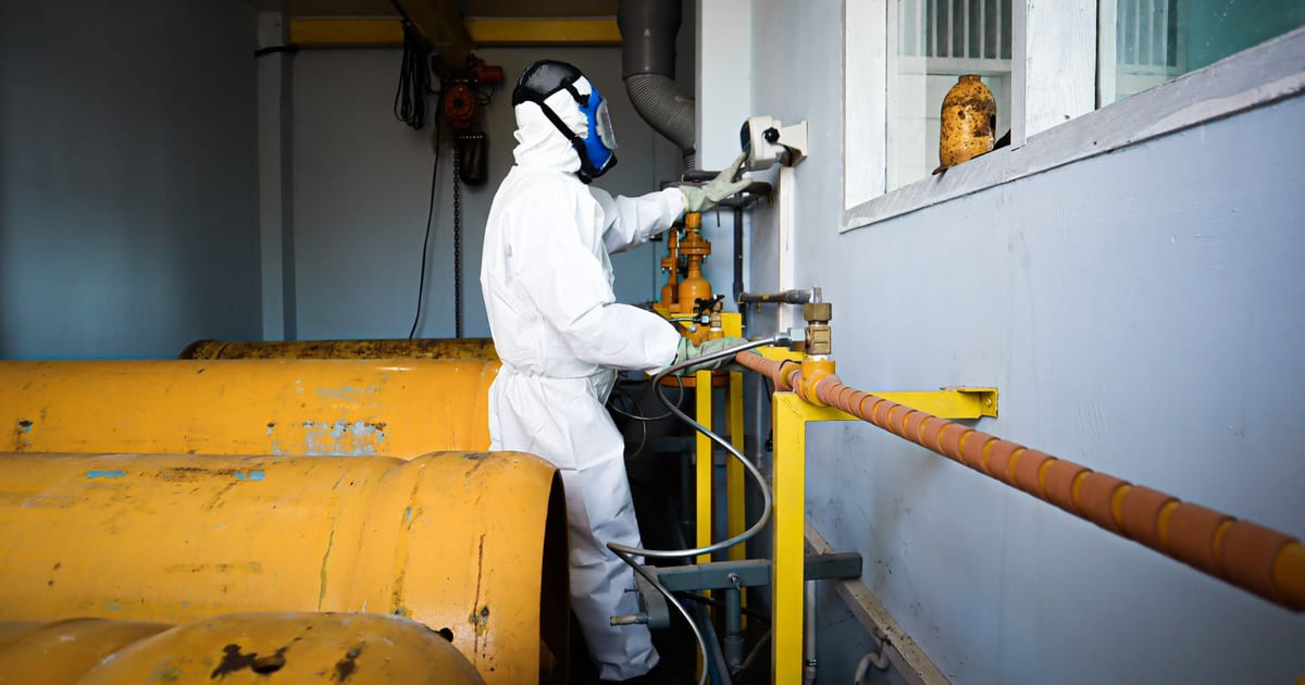 Worker in full protective suit and gas mask handles industrial pipes indoors in Indonesia.
