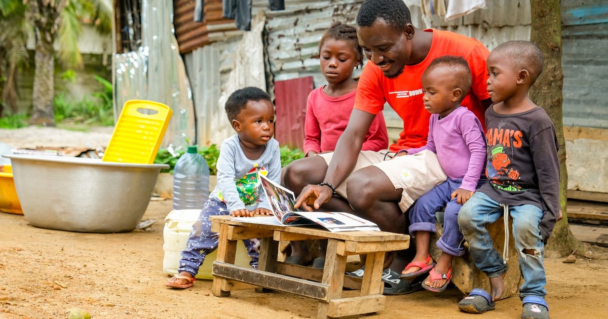 A father reads a book to young children outside a tin house in Monrovia, fostering learning and community.